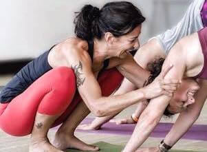 Auf diesem Bild ist Yogalehrerin Andrea, die einer Yogaschülerin im herabschauenden Hund, einer klassischen Yoga Übung, hilft.