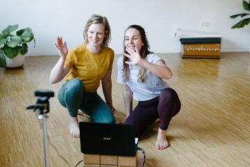 Yohalehrerinnen Christiane und Louisa lächeln und winken in die Laptop-Kamera im Yogastudio Fuß über Kopf in Stuttgart West