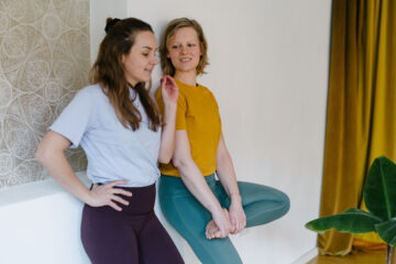 Yogalehrerin Christiane und Yogalehrerin Louisa im Yogastudio Fuß über Kopf. Die beiden unterhalten sich, während sie entspannt an die Wand gelehnt stehen