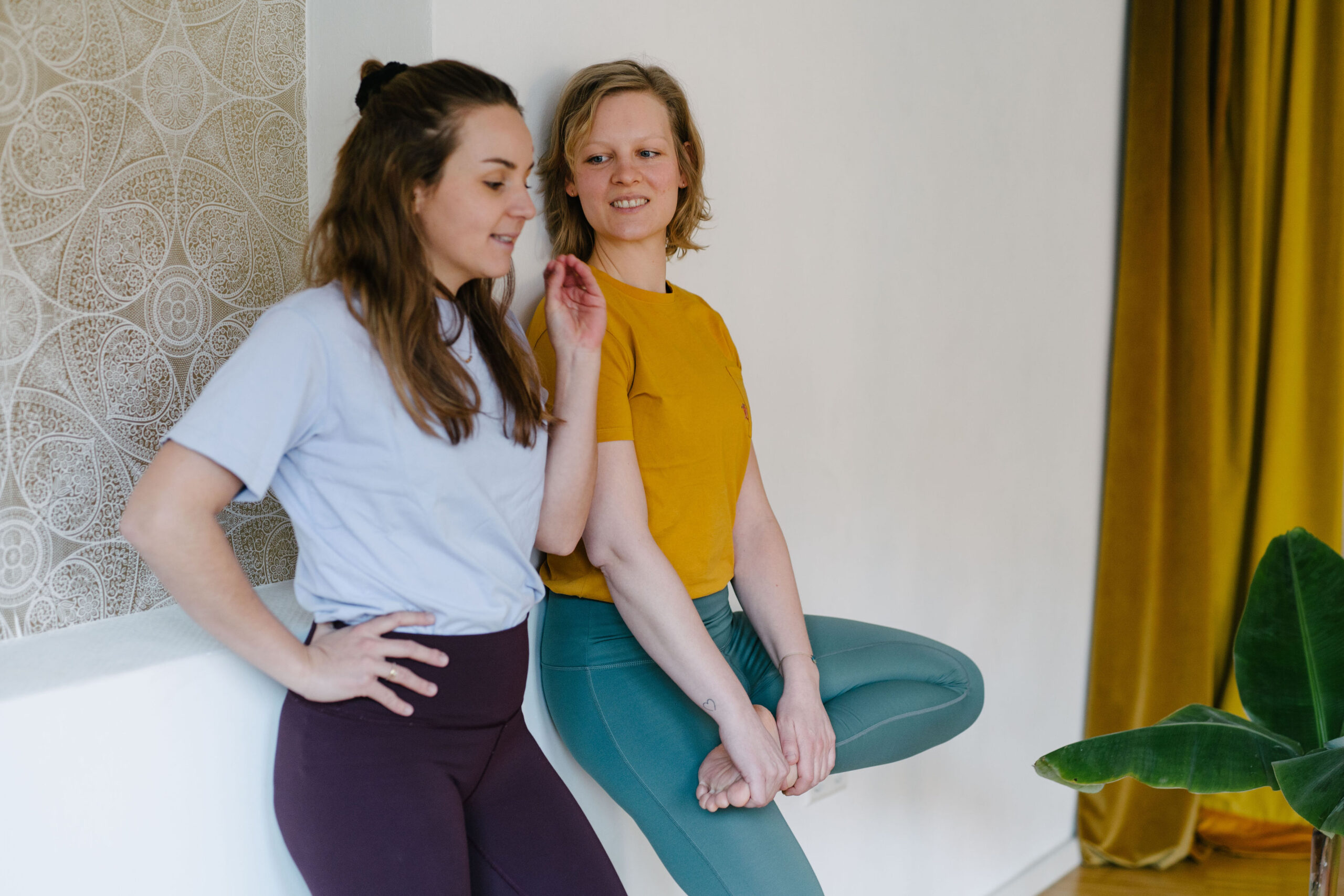 Yogalehrerin Christiane und Yogalehrerin Louisa im Yogastudio Fuß über Kopf. Die beiden unterhalten sich, während sie entspannt an die Wand gelehnt stehen