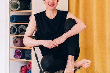 Yogalehrerin Simone Stephan sitzt auf einem Yogastuhl im Yogastudio Fuß über Kopf. Im Hintergrund sind aufgerollte Yogamatten und ein okkerfarbener Vorhang zu sehen.