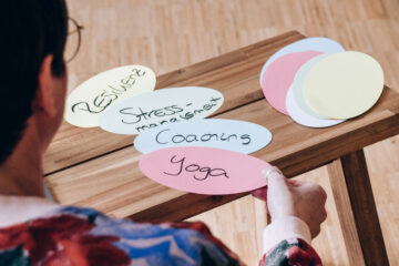Simone Stephan ist Yogalehrerin und Coachin für Stressmanagement und Resilienz. Auf dem Bild hält sie ovale, bunte Karteikarten in der Hand, auf denen Begriffe stehen wie Yoga, Resilienz und Coaching.