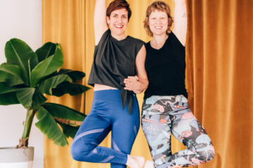 Yogalehrerinnen Simone und Christiane stehen gemeinsam als Partner-Yogaübung im Baum im Yogastudio Fuß über Kopf.